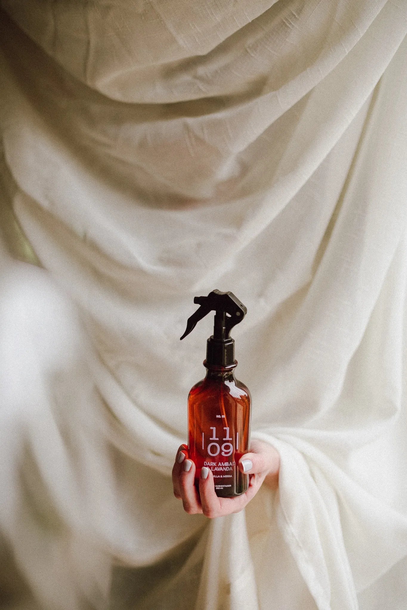 Home spray Dark Ámbar y Lavanda 1109 Concept sostenido en mano con fondo drapeado color marfil